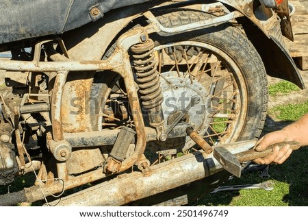 worker using iron wrenches and hammer with wooden handle by hand removes rear dirty rusty old rubber tire and spokes wheel from retro heavy motorcycle on green grass during daytime outdoors