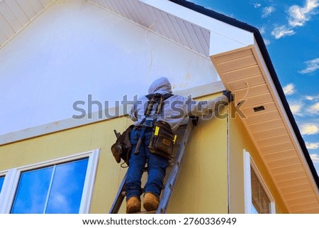 Worker is using ladder to fix trim on house while installing fiber siding panels