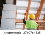 Worker using level tool adjusts wooden rafter during roofing, brick wall backdrop, house structure at roof installation stage.