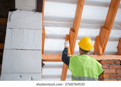 Worker using level tool adjusts wooden rafter during roofing, brick wall backdrop, house structure at roof installation stage.