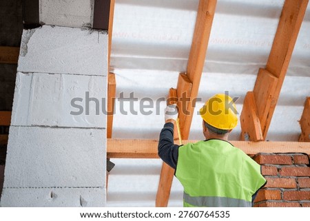 Worker using level tool adjusts wooden rafter during roofing, brick wall backdrop, house structure at roof installation stage.