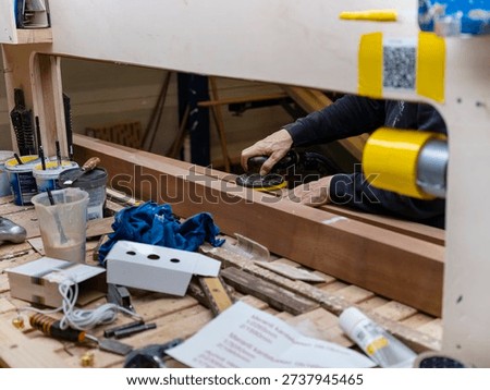 Worker using orbital sander on wooden doorframe for finishing surface preparation in factory workshop setting