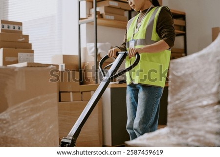 Worker using a pallet jack to move boxes in a warehouse surrounded by shelves and storage units wearing reflective safety vest moving heavy packages
