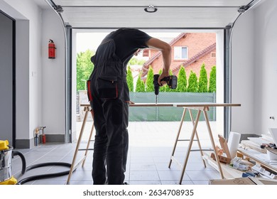Worker using power drill on wooden board supported by sawhorses in home garage workshop. Man working in workshop, drilling hole in chipboard for furniture assembly
