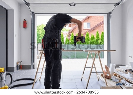 Worker using power drill on wooden board supported by sawhorses in home garage workshop. Man working in workshop, drilling hole in chipboard for furniture assembly