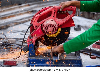 Worker using a red abrasive cut-off saw to cut metal at a construction site. Bright orange sparks flying from the blade as it slices through a steel bar in a workshop environment.