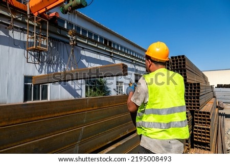 Worker using remote control of gantry crane to transport stack of metal pipes in the steel factory