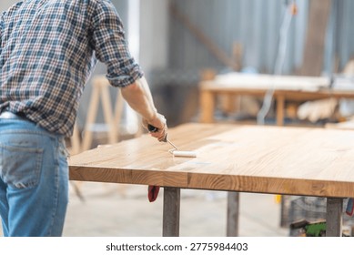 Worker using a roller to apply wood finish on a large oak table in a workshop