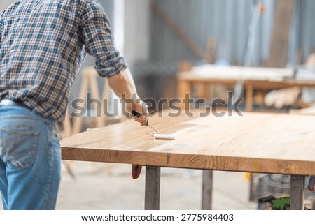 Worker using a roller to apply wood finish on a large oak table in a workshop