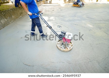 Worker using self-leveling power trowel machine with gas engine has wheels with pads, plate and he is driving it in circles smoothing surface on concrete slab.