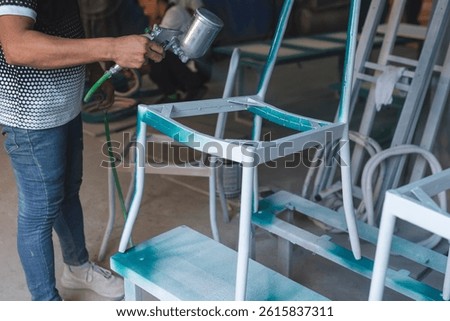 Worker using a spray gun to apply teal paint on a steel or iron chair frame during furniture finishing in a production workshop.