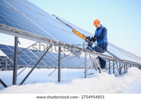 Worker using tools on solar panels covered in snow, performing maintenance at a renewable energy farm during winter