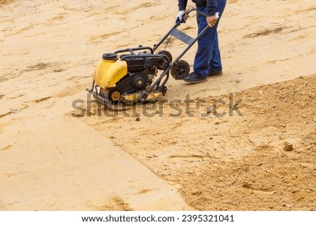 Worker using vibratory plate compactor for compaction sand during path construction at summer day.