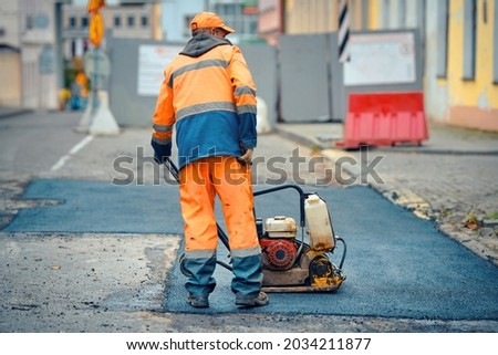 Worker with vibratory plate compactor for road construction. Road repair, worker paving fresh bitumen. Pothole repair, asphalt paving, patching road works with plate compactor.