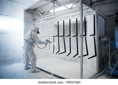 Worker wearing protective wear performing powder coating of metal details in a special industrial camera