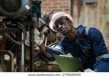 Worker wears a white hard hat and blue work jacket. He uses safety glasses while handling industrial machinery. Clipboard rests in his hand. Brick walls form the workshop background