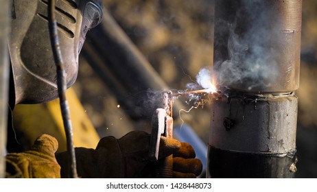 Worker Is Welding Steel Pipes Outdoors In Daytime On An Oil Well, Wearing Protective Mask And Gloves