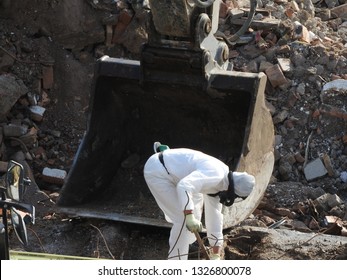 Worker With White Protective Suit, Gloves And Mask Removes Removal White Asbestos On Construction Site. Demolition Building With Excavator. Action Picture, Part Of A Serie.