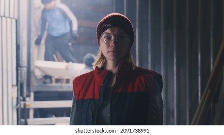 Worker Woman In Vest And Hat Looking At Camera Against Entrance Of Workshop While Colleagues Unloading Truck With Materials