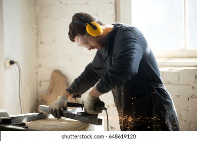 Worker In Workwear, Protective Glasses, Hearing Protection Headphones, Gloves Using Dangerous Electric Power Tools In Loft Workshop. Safety At Workers Workplace, Personal Protective Equipment Concept