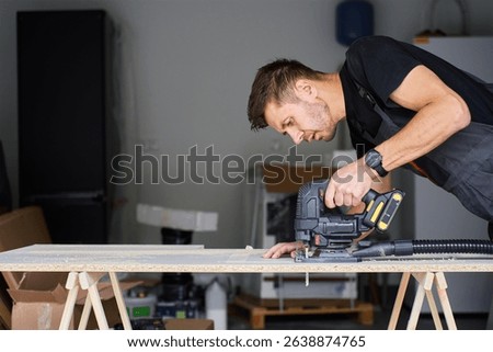 Worker in workwear using handheld cordless electric jigsaw to cut wooden board in garage. Skilled carpenter working in workshop. Man doing DIY home renovation