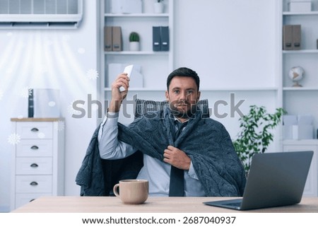 Worker wrapped in blanket using remote control at table in office. Broken or incorrectly adjusted air conditioner