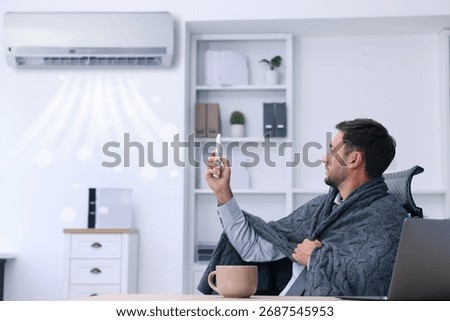 Worker wrapped in blanket using remote control at table in office. Broken or incorrectly adjusted air conditioner
