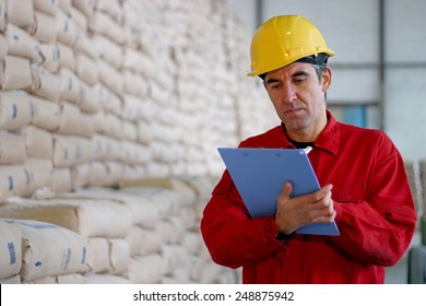 Worker Writing On Clipboard In Warehouse. Warehouse Worker At Work. Worker In Distribution Warehouse Beside Big Pile Of Sacks.