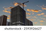 Workers are actively constructing a tall building, utilizing a crane to lift materials as the sun sets, casting a warm glow over the site and surrounding structures.
