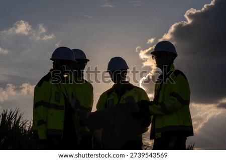 Workers analyze plans under the sunset at a construction site while wearing safety gear and helmets in the evening sky