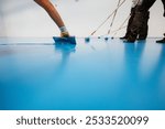 Workers applying a blue epoxy resin on a floor with rollers and spatulas in an industrial setting.