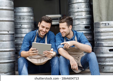 Workers Brewers Managers And Modern Technology For Business Management. Smiling Young Male Workers In Aprons Sit, Gesturing And Look At Data In Tablet In Interior Of Warehouse With Metal Kegs