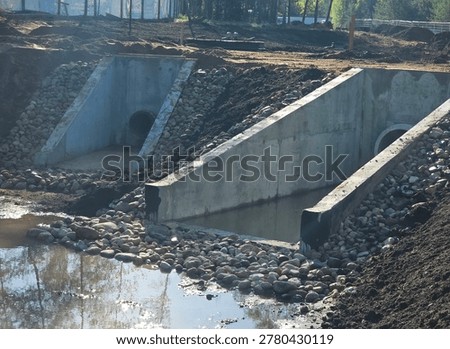 Workers are building drainage channels in a construction area. Two concrete structures connect to the water. Stones surround the openings and water flows through them.
