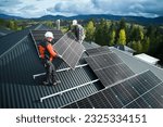 Workers building solar panel system on roof of house. Men technicians in helmets carrying photovoltaic solar module outdoors. Concept of alternative and renewable energy.