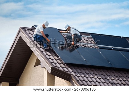 Workers building solar panel system on rooftop of house. Two men installers in helmets installing photovoltaic solar module outdoors. Alternative, green and renewable energy generation concept.