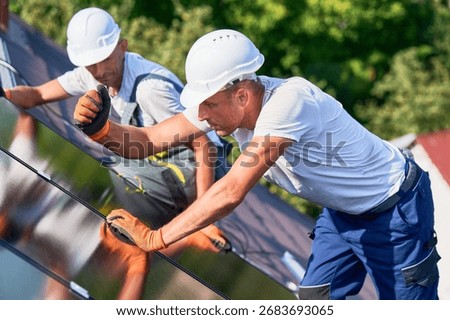Workers building solar panel system on rooftop of house. Two men installers in helmets and gloves installing photovoltaic solar module outdoors. Alternative and renewable energy generation concept.