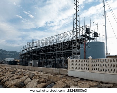 Workers are busy at a boatyard refit facility located by the coast. Scaffolding supports renovations on boats with water storage equipment seen nearby. Stone breakwater rocks line the shore.