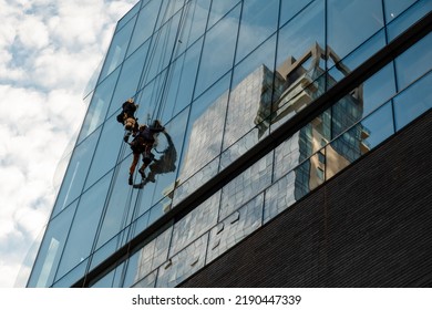 Workers Cleaning Office Building Windows. Industrial Climbers Wash The Windows Of A Modern Skyscraper. Men Wash Windows On The Evolution Tower. Washers Wash Skyscraper Windows Sitting On Hanging