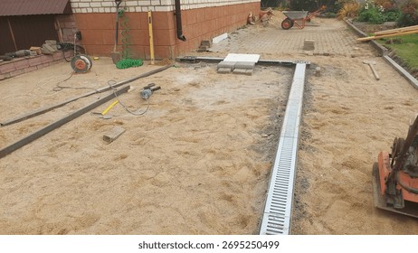 Workers are constructing a drainage system in a backyard. Various construction tools and materials are visible, and the ground is covered with sand, indicating ongoing work.