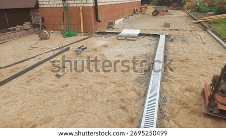 Workers are constructing a drainage system in a backyard. Various construction tools and materials are visible, and the ground is covered with sand, indicating ongoing work.