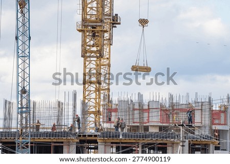 Workers developing a new high-rise building on a busy construction site, with a large tower crane lifting a bundle of wooden planks against a cloudy sky, symbolizing urban expansion