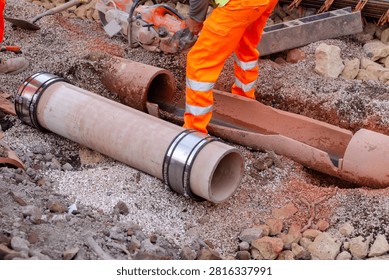 Workers dig in the ground to place a large pipe. Tools and materials are scattered around. The scene shows construction activity in an urban area.