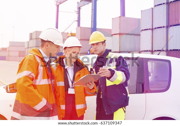 Workers discussing over clipboard beside car in\
shipping yard