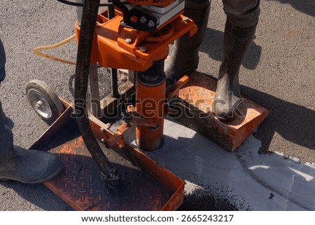 Workers drilling asphalt core sample with orange coring machine during inspection.