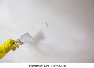 Worker's Hand In Yellow Gloves Holding Putty Knife Patching A Hole With Spatula With Plaster Or Putty In White Wall. Renovation And Repair Process, Remodeling Interior Of Room At Apartment Building