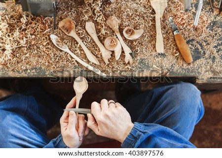 Worker's hands in blue jeans working suit carving a wooden spoon with a knife, shavings on table at background, close up, woodworking, copy space.