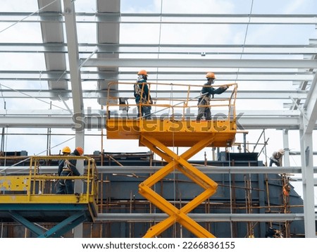 Workers install lighting fixture in a hug industrial warehouse using hydraulic scissor lift. MEP work in a construction site.