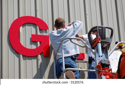 Workers install a sign on the side of an industrial building.
