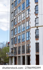 Workers installing facade system and thermal insulation on unfinished apartment building. Modern residential construction site with suspended platforms and exposed concrete structure.