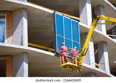 Workers in lift bucket installing large glass panes on new building at construction site. Workers team in cradle installing glass window on building. Installation of insulated double-glazed windows. 